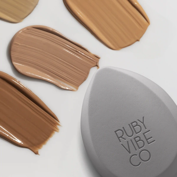 A grey Ruby Vibe Co makeup sponge rests beside four different shades of liquid foundation swatches.